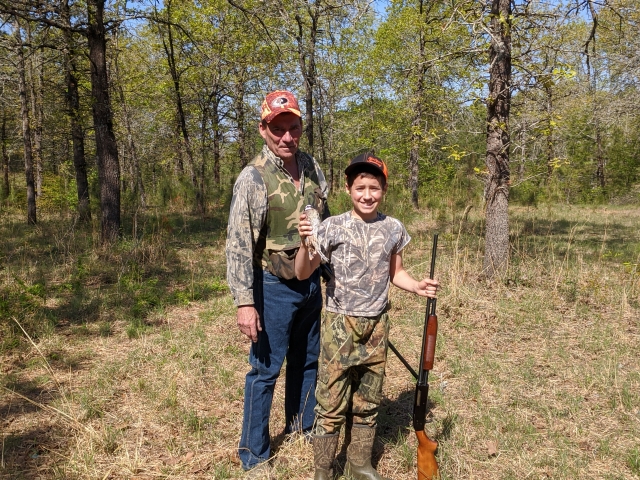 A man and a boy standing in a wooded area with a rifle.