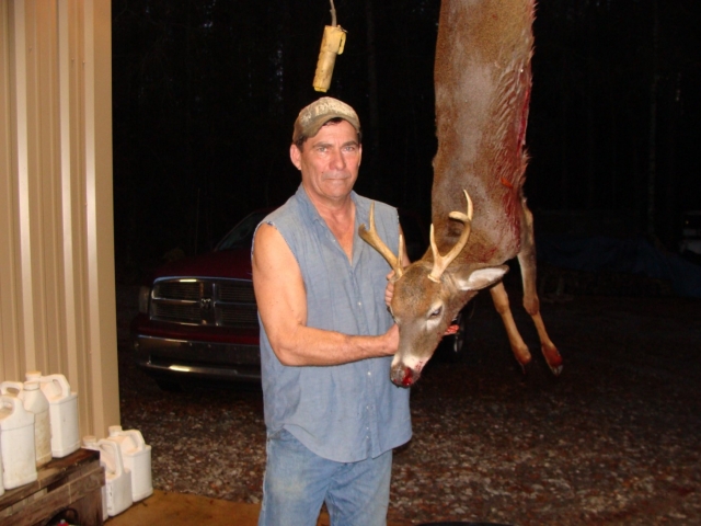 A man holding a dead deer in front of a barn.