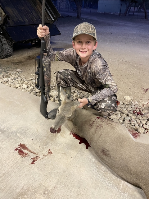 A young boy posing with a rifle next to a dead deer.