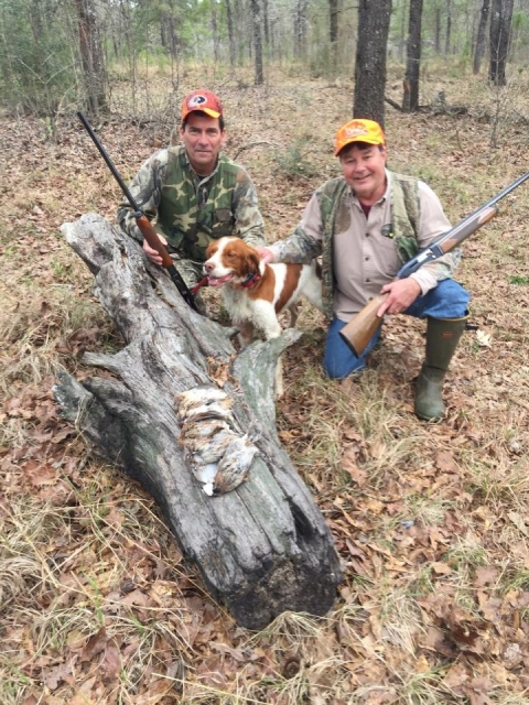 Two men and a dog posing next to a log in the woods.